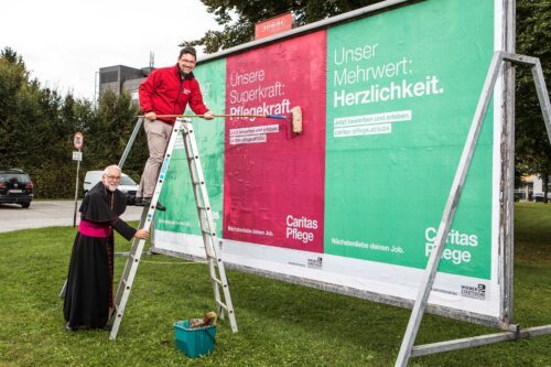 Caritasdirektor Ernst Sandriesser und sein Vorgänger Bischof Josef Marketz haben sich mit Leiter, Kleister und großflächigen Plakaten auf den Weg gemacht, um Verstärkung zu suchen. Foto – © Caritas/gleissfoto